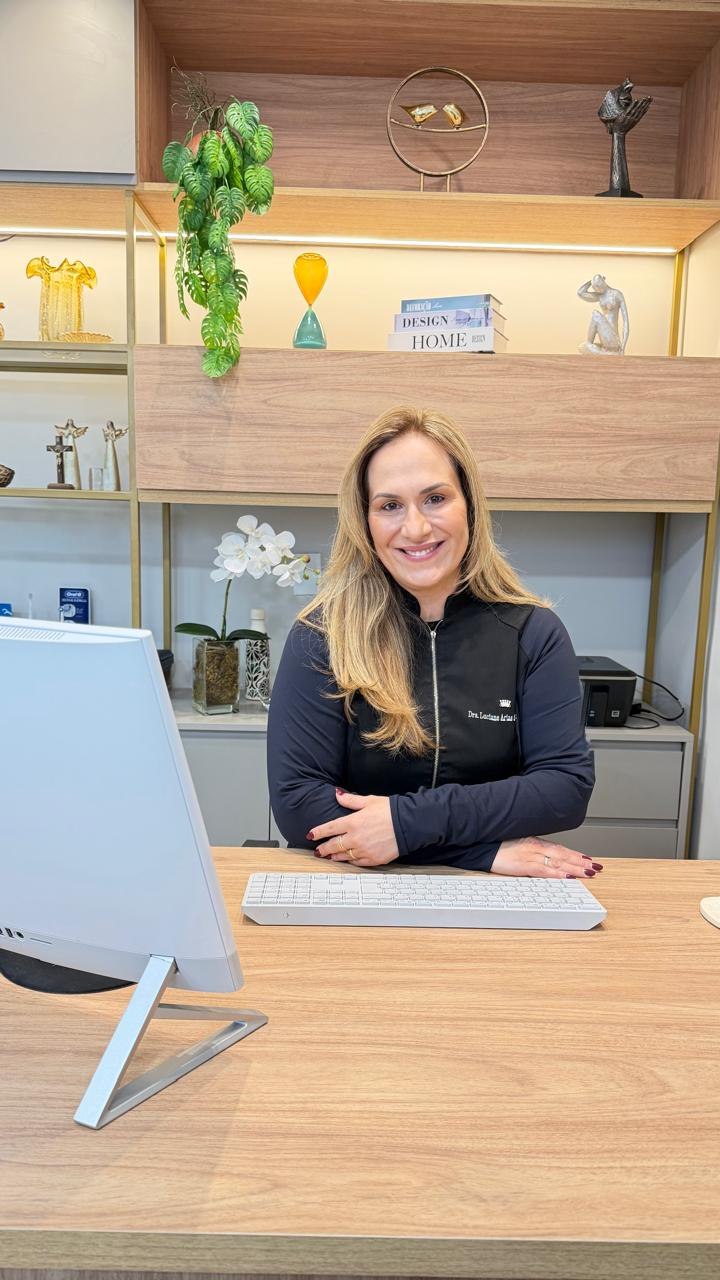 Dra. Luciane Arias em seu consultório, com plantas e uma estante iluminada ao fundo